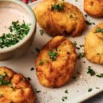 Five breaded and fried potato croquettes garnished with chopped parsley are arranged on a plate next to a bowl of dipping sauce topped with herbs.