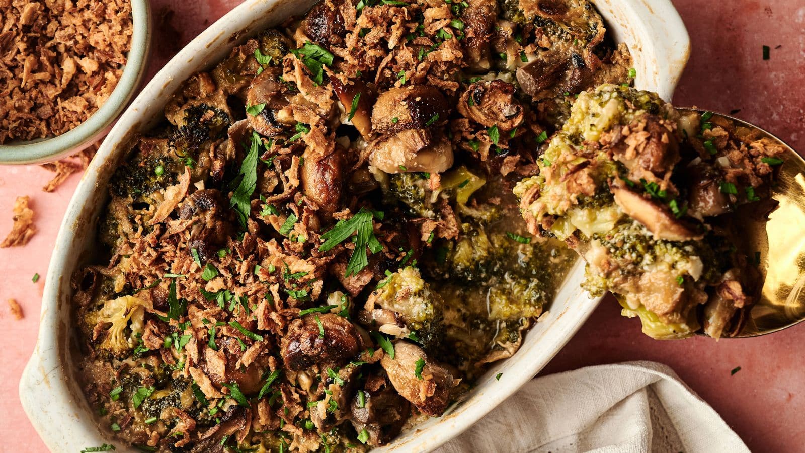 A casserole dish filled with baked broccoli and mushrooms topped with crispy fried onions, with a serving spoon lifting out a portion.