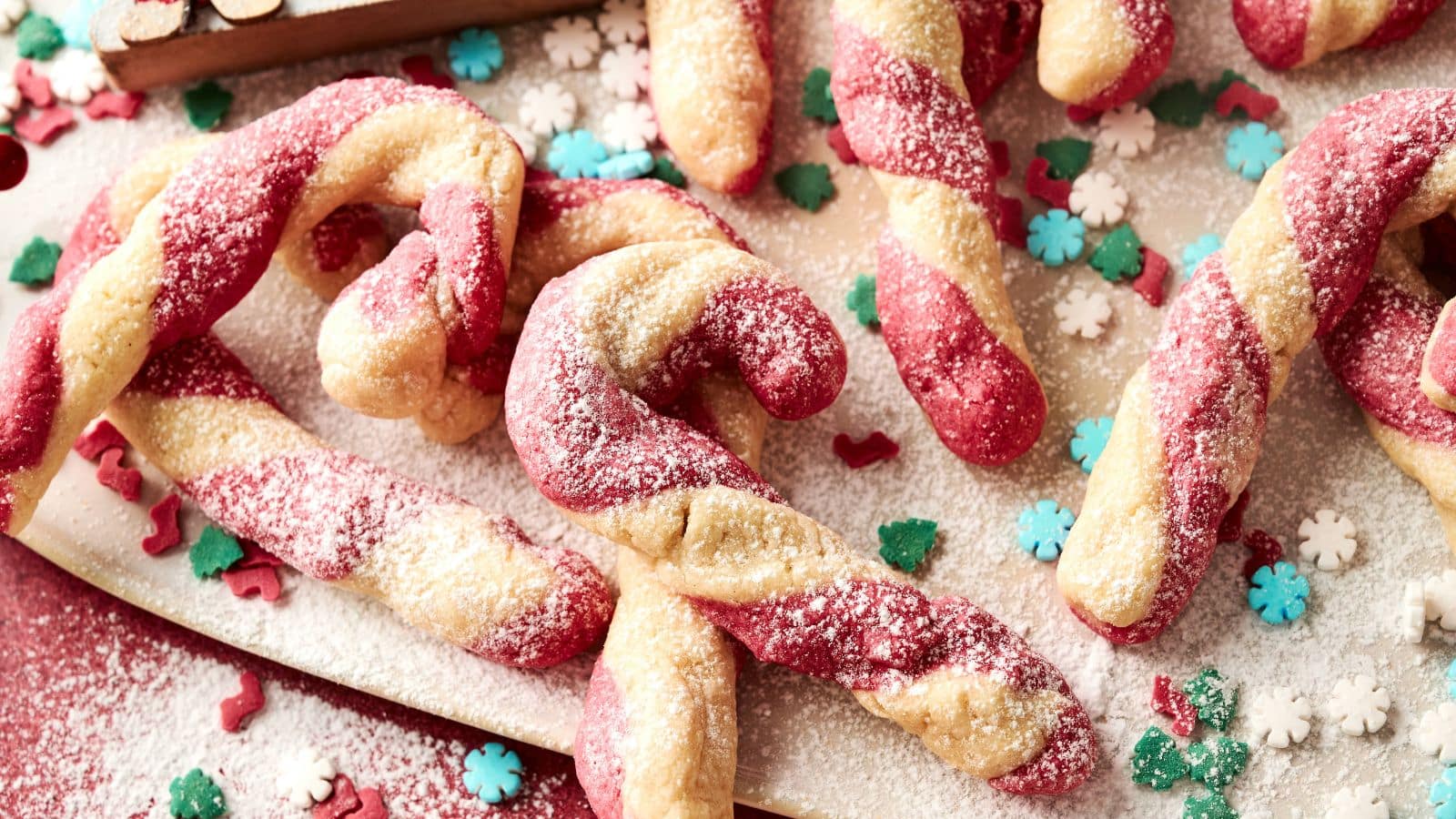 Twisted red and white candy cane cookies dusted with powdered sugar, surrounded by decorative snowflake and tree-shaped sprinkles.