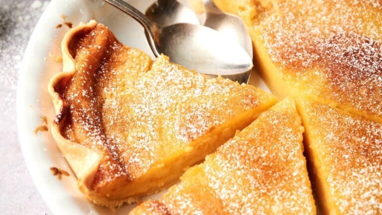 A close-up of a lemon tart cut into slices, dusted with powdered sugar, with two metal spoons resting on the pie plate.