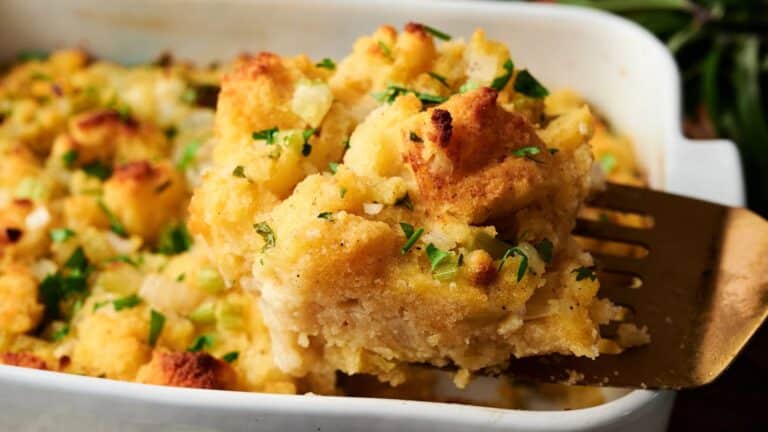 A serving spatula lifts a portion of baked bread stuffing with herbs from a white casserole dish.