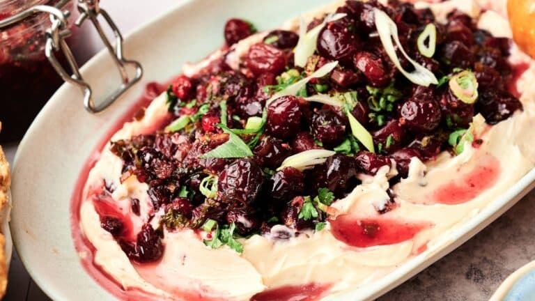 A serving dish with whipped cheese topped with roasted cranberries, herbs, and sliced green onions.