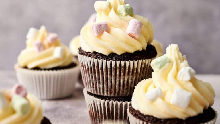 Chocolate cupcakes with white frosting are topped with mini pastel-colored marshmallows, arranged on a light surface with a neutral background.