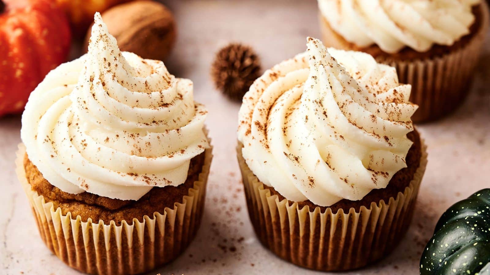 Two cupcakes with white swirled frosting and a sprinkle of cinnamon sit on a light surface, surrounded by fall decorations.