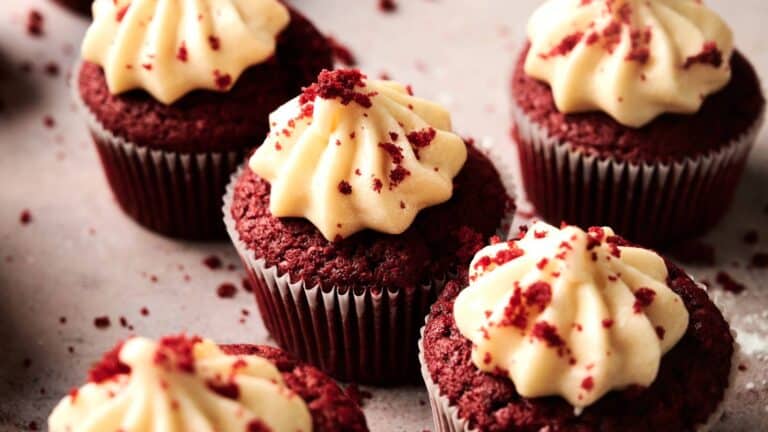 Close-up of several red velvet cupcakes topped with swirls of cream cheese frosting and red cake crumbs, arranged on a light surface.