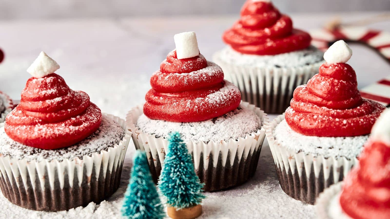 Chocolate cupcakes with red frosting swirled to resemble Santa hats, topped with powdered sugar and a small marshmallow, with miniature decorative trees in the foreground.