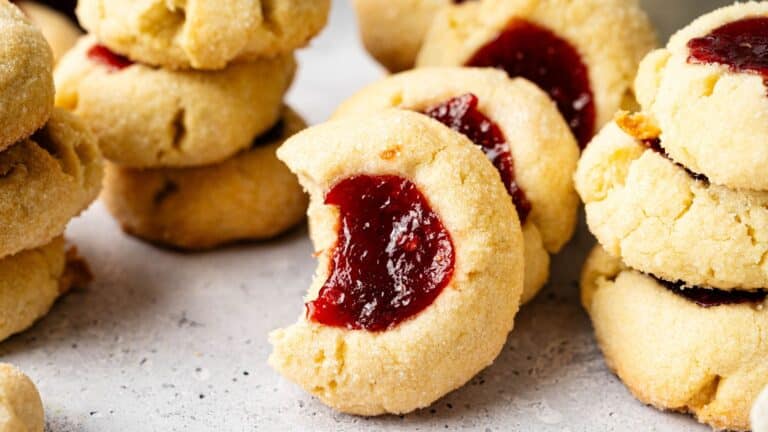 Close-up of several thumbprint cookies filled with red jam, with one cookie showing a bite taken out of it.