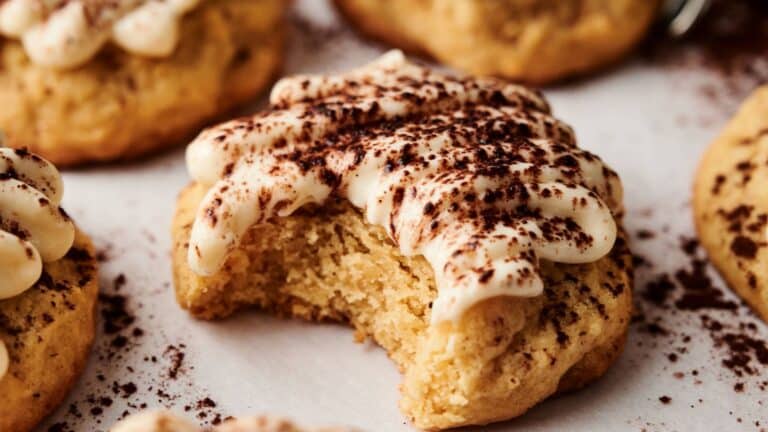 A soft cookie with white icing and cocoa powder on top, shown with a bite taken out, sits on parchment paper among other similar cookies.