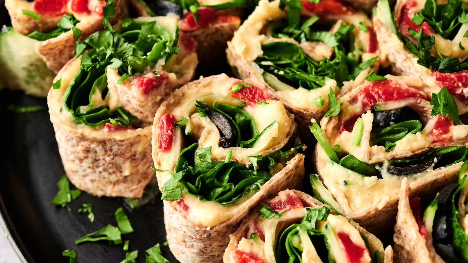 Close-up of sliced veggie pinwheels made with tortilla wraps, hummus, spinach, roasted red peppers, olives, and fresh herbs arranged on a plate.