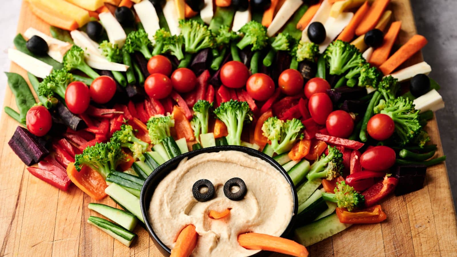 A colorful assortment of fresh vegetables arranged like a turkey on a wooden board, with a bowl of hummus decorated as a face using olives and a carrot.