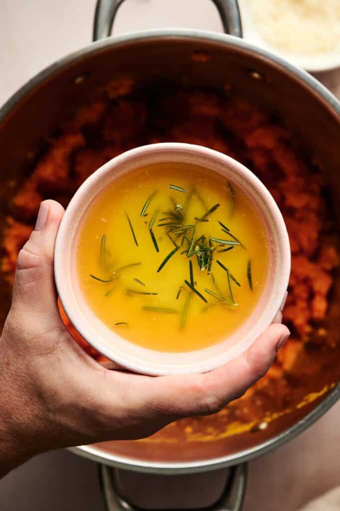 A hand holds a small bowl of melted butter garnished with rosemary over a pot filled with creamy Mashed Sweet Potatoes.