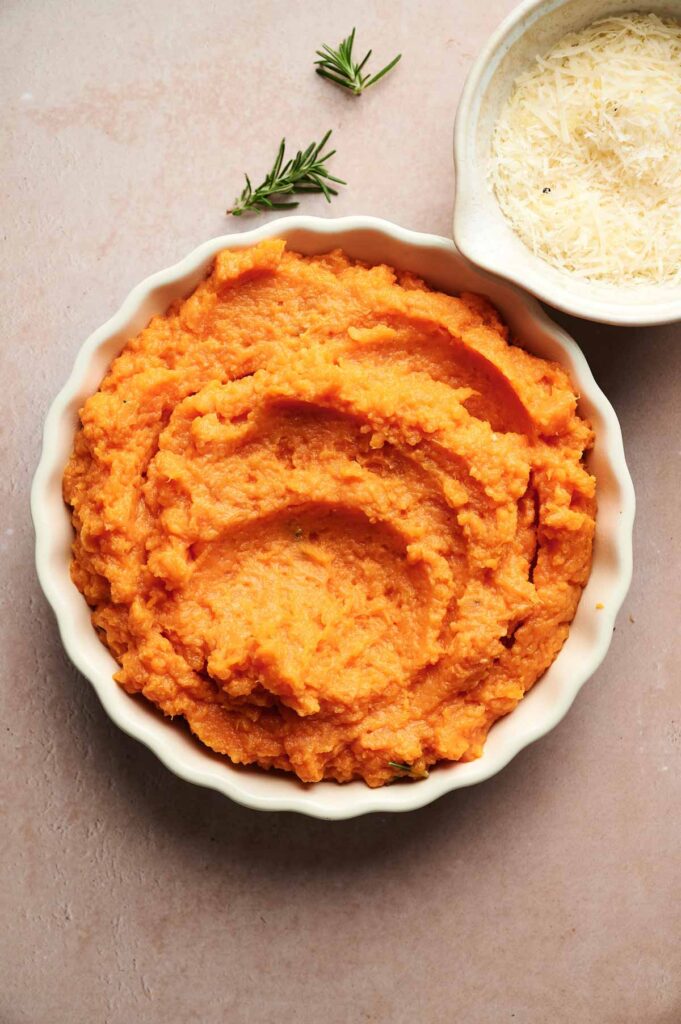 A round dish of Mashed Sweet Potatoes sits next to a small bowl of grated cheese and two sprigs of rosemary on a light countertop.
