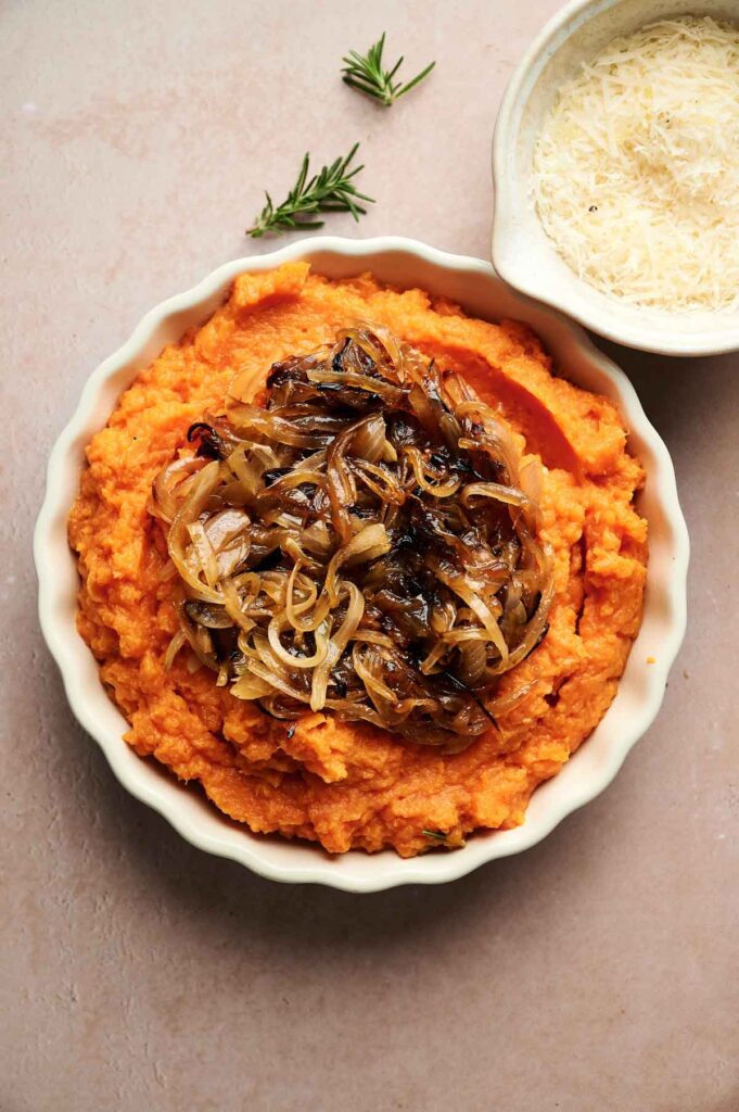A comforting dish of Mashed Sweet Potatoes topped with caramelized onions, served alongside a small bowl of shredded cheese and a sprig of rosemary for garnish.