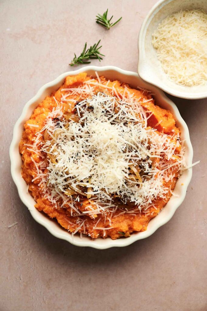 A round dish of Mashed Sweet Potatoes topped with grated cheese, served with a small bowl of extra cheese and fresh rosemary sprigs on the side.