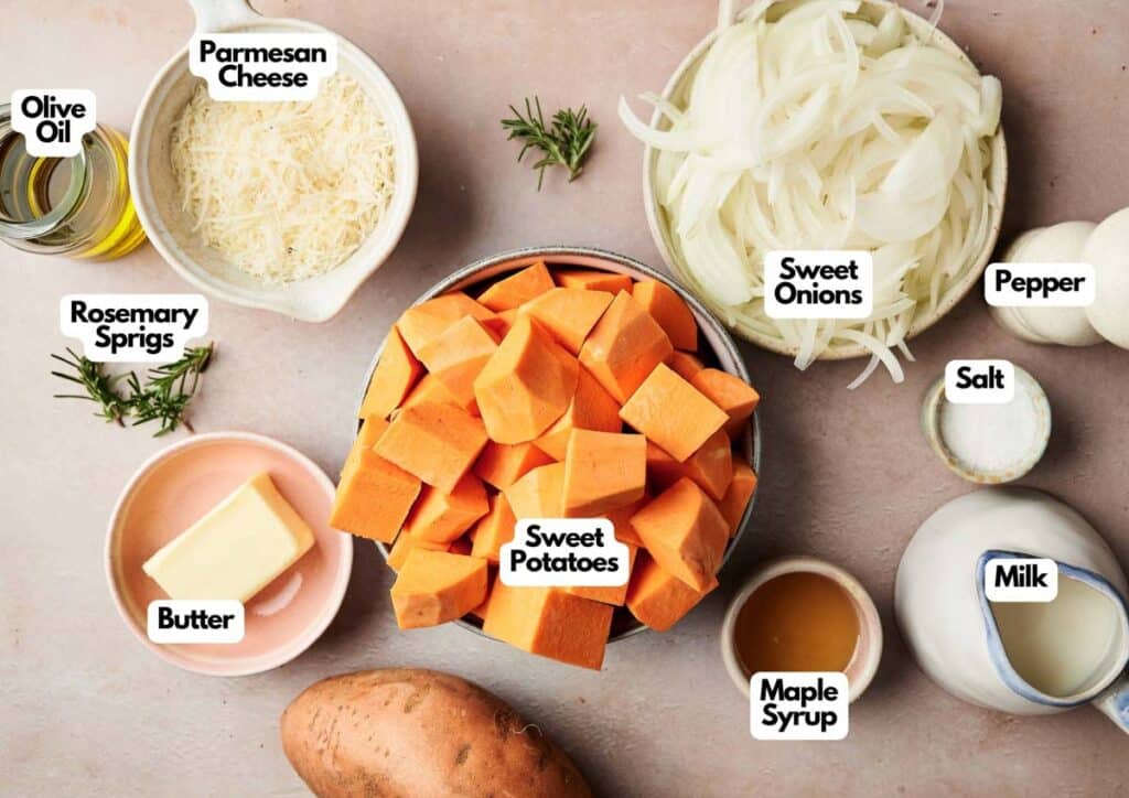 Assorted ingredients for Mashed Sweet Potatoes on a table, including cubed sweet potatoes, sliced sweet onions, grated parmesan cheese, butter, olive oil, rosemary, maple syrup, milk, salt, and pepper.