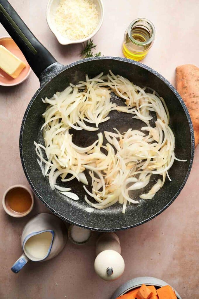 Sliced onions cooking in a black frying pan, surrounded by small bowls of butter, oil, grated cheese, cream, and mashed sweet potatoes on a light countertop.