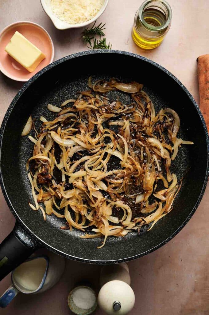 A skillet with caramelized onions sits on a counter, surrounded by butter, grated cheese, olive oil, and other ingredients ready for mashed sweet potatoes.