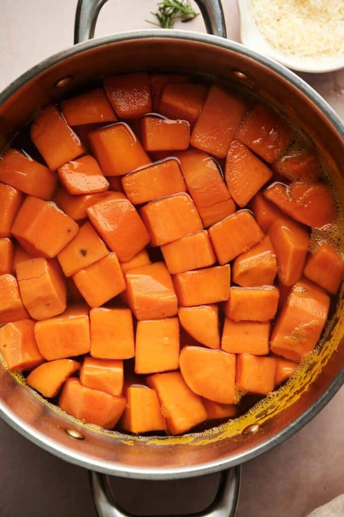 Cubed sweet potatoes simmering in a pot of water, perfect for making creamy Mashed Sweet Potatoes, with a bowl of grated cheese and a sprig of rosemary nearby.