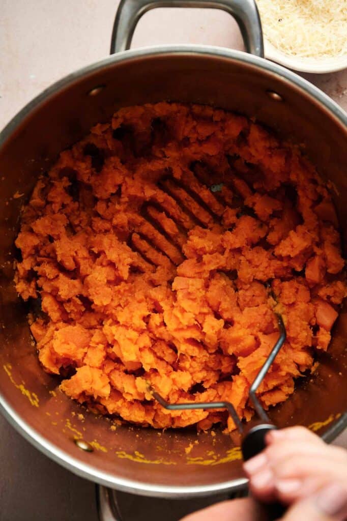 A hand mashing cooked sweet potatoes with a potato masher in a stainless steel pot, creating creamy mashed sweet potatoes.