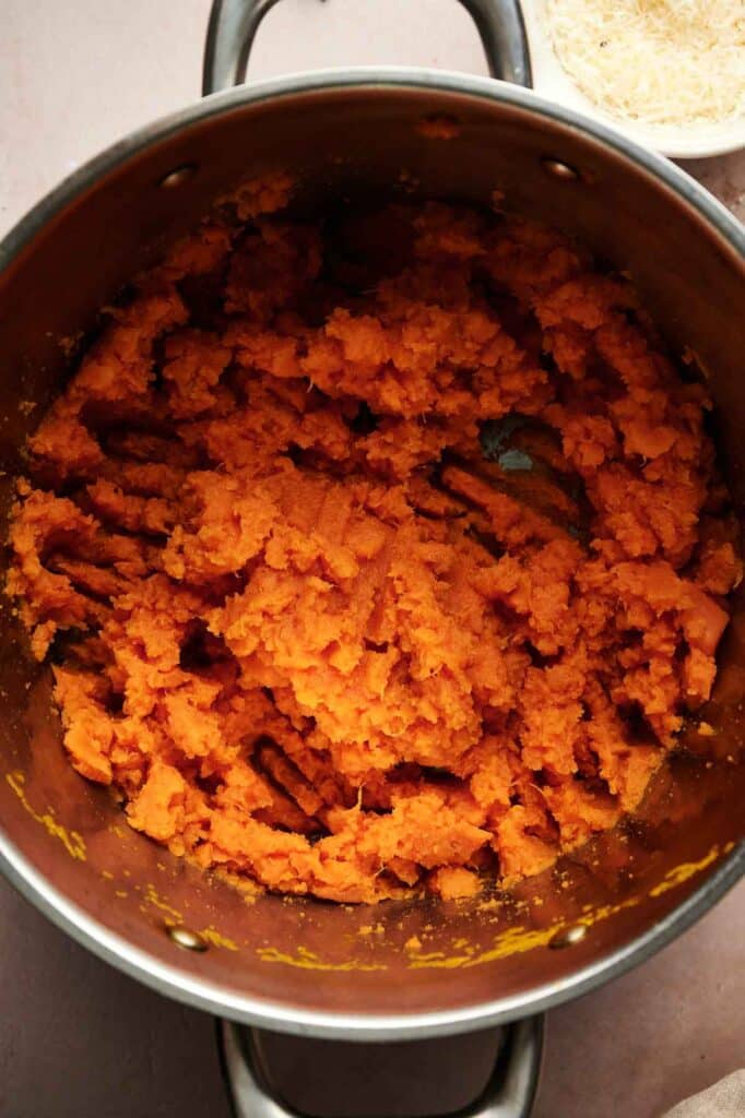 A pot filled with creamy Mashed Sweet Potatoes sits on a light-colored surface, with a dish of shredded cheese partially visible in the upper right corner.