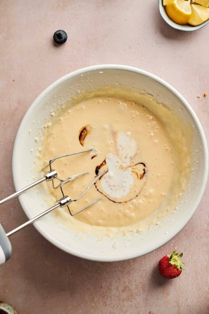 A mixing bowl filled with batter for mini cheesecakes is being blended with a hand mixer; vanilla and milk are visible on the surface. Fruit and lemon wedges are on the table nearby.