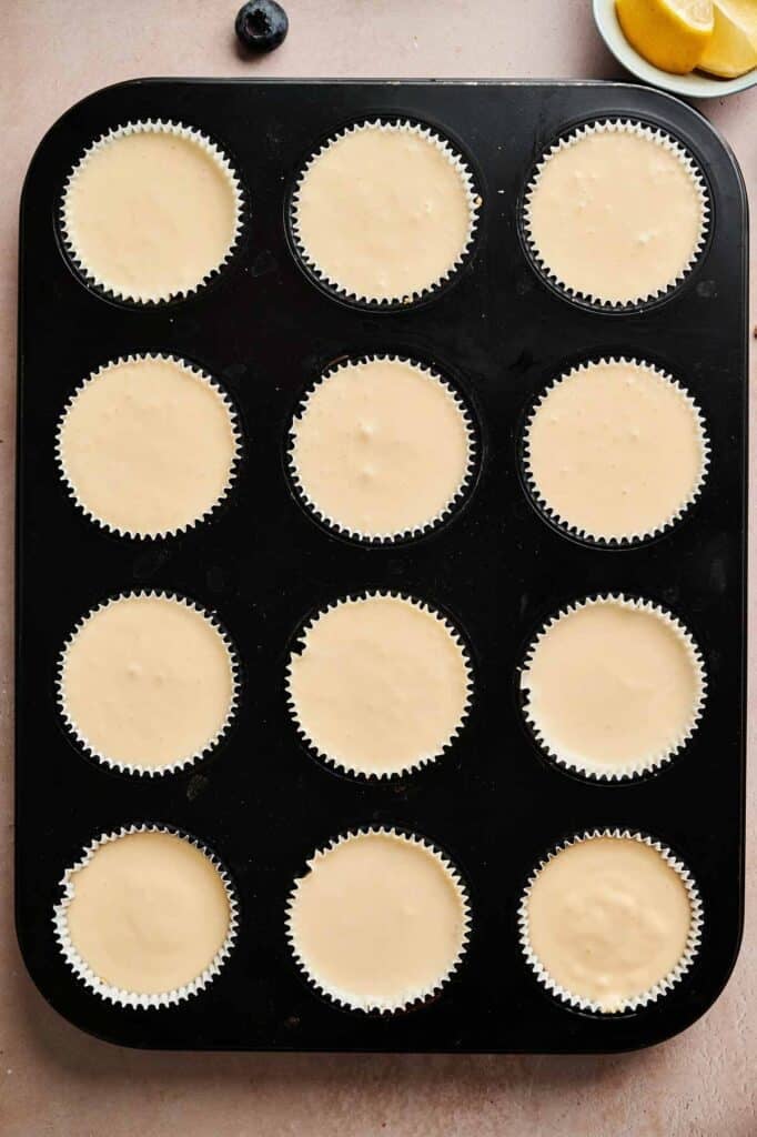 A muffin tin with twelve paper liners filled with pale, unbaked Mini Cheesecakes batter, ready to be baked. Lemon slices and a blueberry are visible in the background.