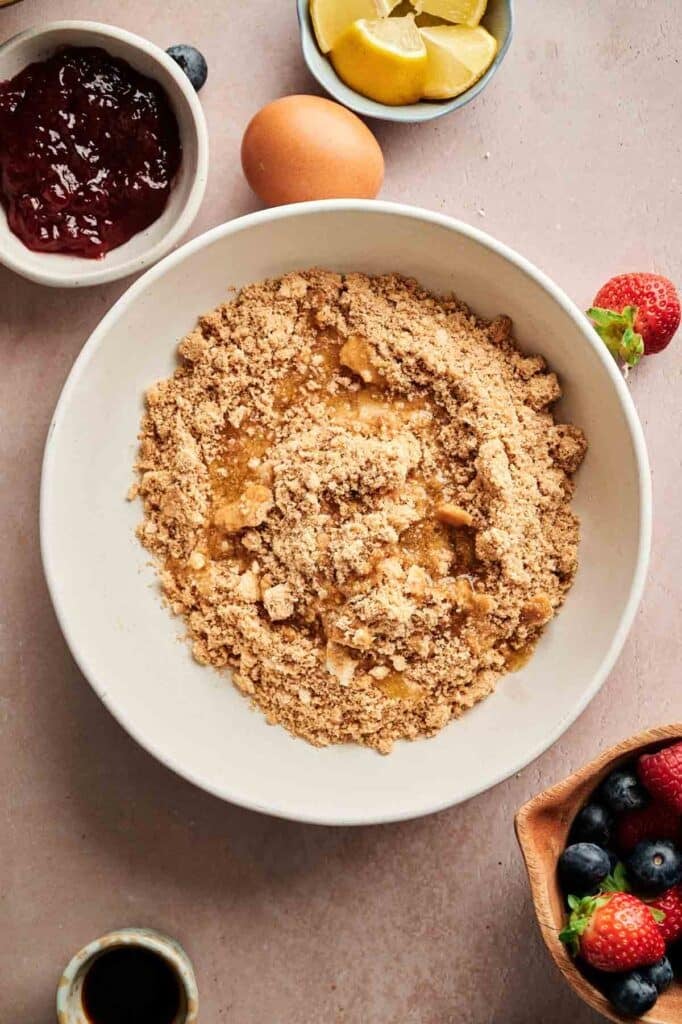 A bowl of crumbly flour mixture, perfect for making Mini Cheesecakes, is surrounded by an egg, lemon wedges, a bowl of jam, fresh berries, and a small cup of liquid.