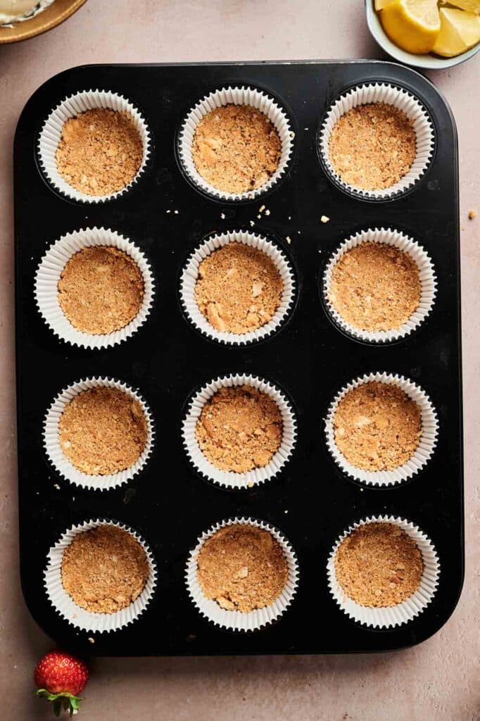 A muffin tin lined with white paper cups, each filled with pressed graham cracker crust for mini cheesecakes, sits on a light countertop.