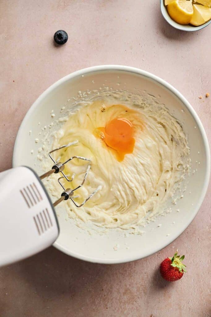 An electric hand mixer blends batter for mini cheesecakes in a white bowl with an egg on top; fresh strawberry, blueberry, and lemon slices rest nearby on a light countertop.