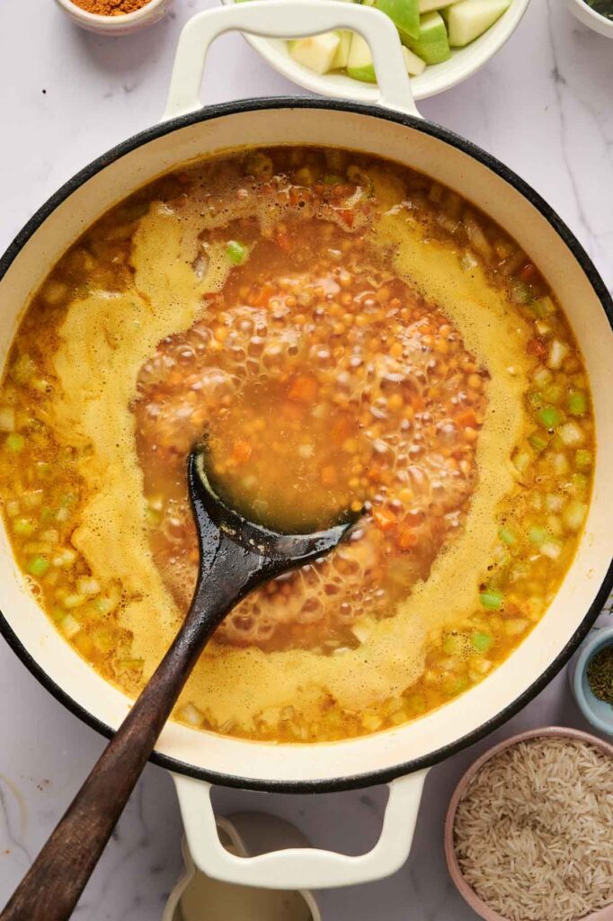 A pot of Mulligatawny Soup with vegetables and broth is being stirred with a wooden spoon; bowls of ingredients surround the pot.