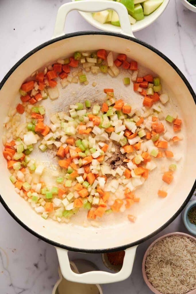Chopped onions, carrots, and celery are sautéing in a large white pot on a marble countertop, as the base for Mulligatawny Soup, with bowls of uncooked rice and vegetables nearby.