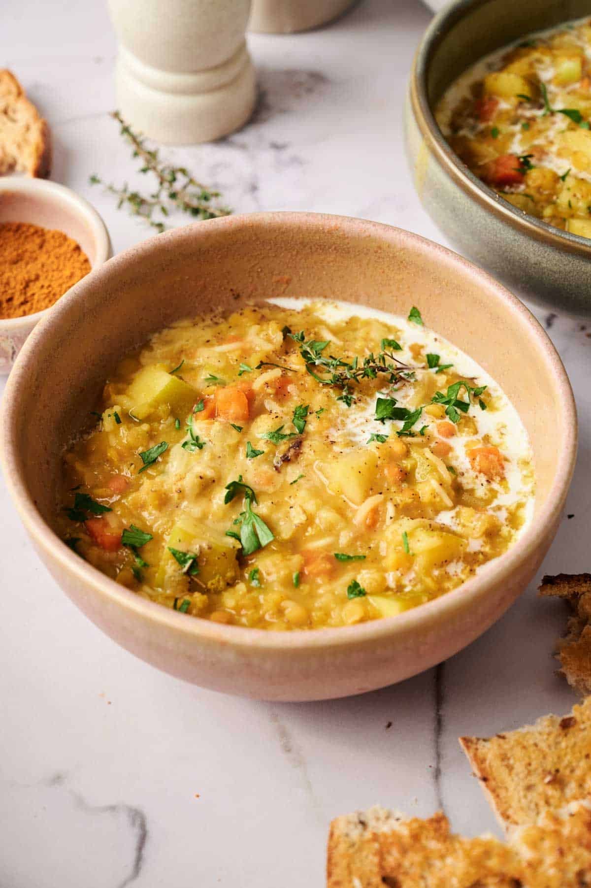 A bowl of creamy Mulligatawny Soup garnished with fresh herbs sits on a marble surface, accompanied by bread slices and a small dish of spice nearby.