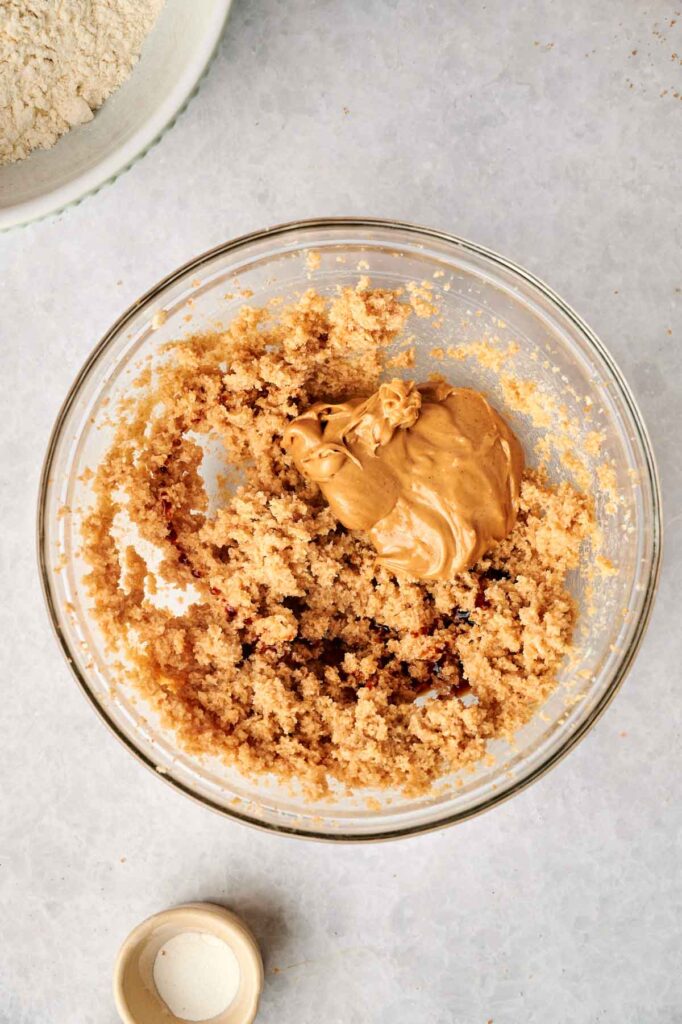 A glass bowl containing a mixture of creamed sugar and butter with a dollop of peanut butter on top, set on a light countertop—perfect for making delicious Air Fryer Peanut Butter Cookies, with a bowl of flour and a small dish nearby.