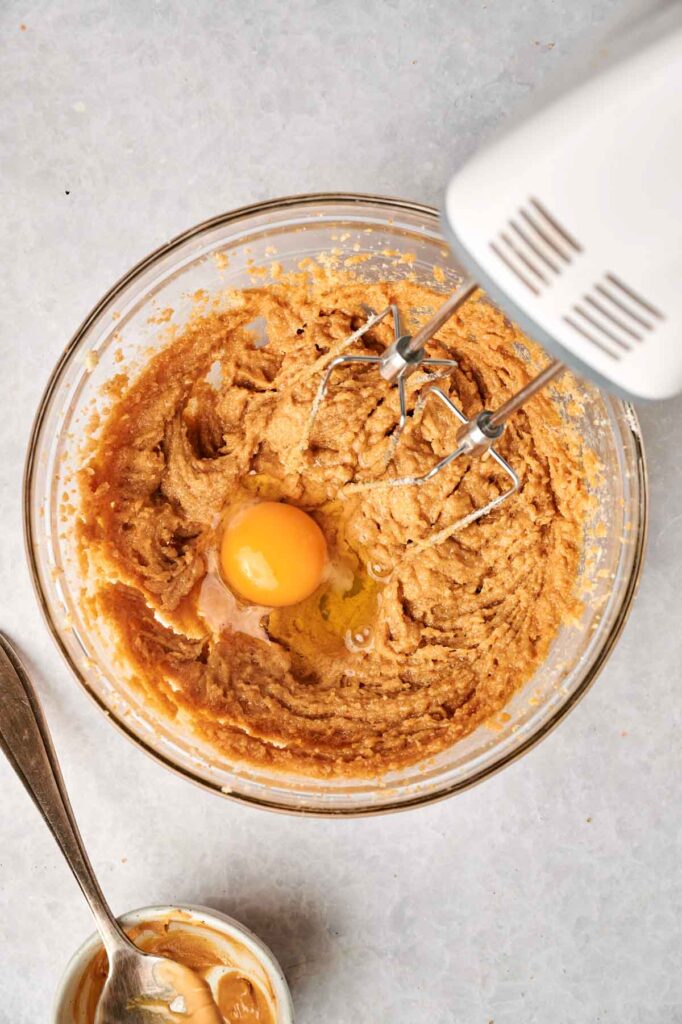 A mixing bowl with cookie dough for Air Fryer Peanut Butter Cookies, an egg on top, and electric mixer beaters partially submerged, with a small dish and spoon beside it on a light surface.
