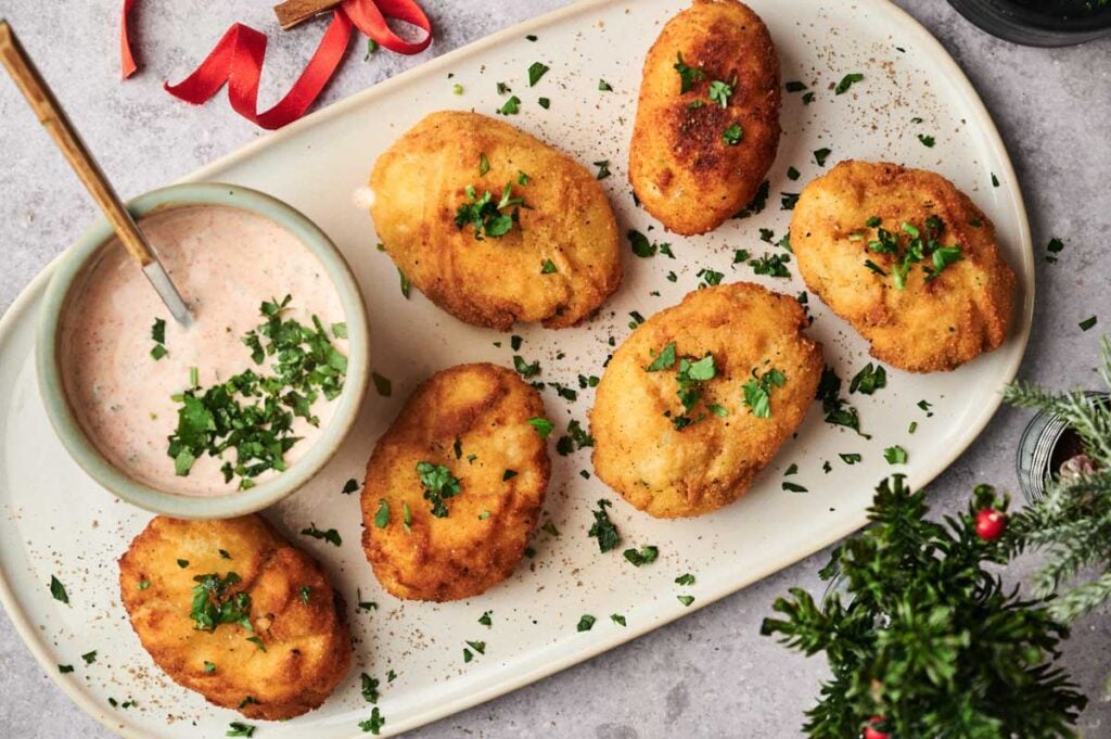 Six breaded potato croquettes garnished with chopped parsley are served on a platter with a small bowl of creamy dipping sauce, sprinkled with herbs.
