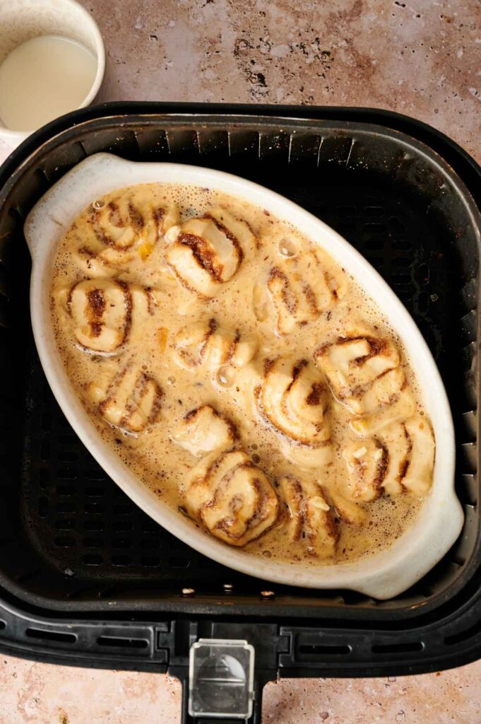 An oval dish filled with unbaked Air Fryer Cinnamon Roll Casserole, topped with creamy sauce, sits inside an air fryer basket on a mottled countertop.