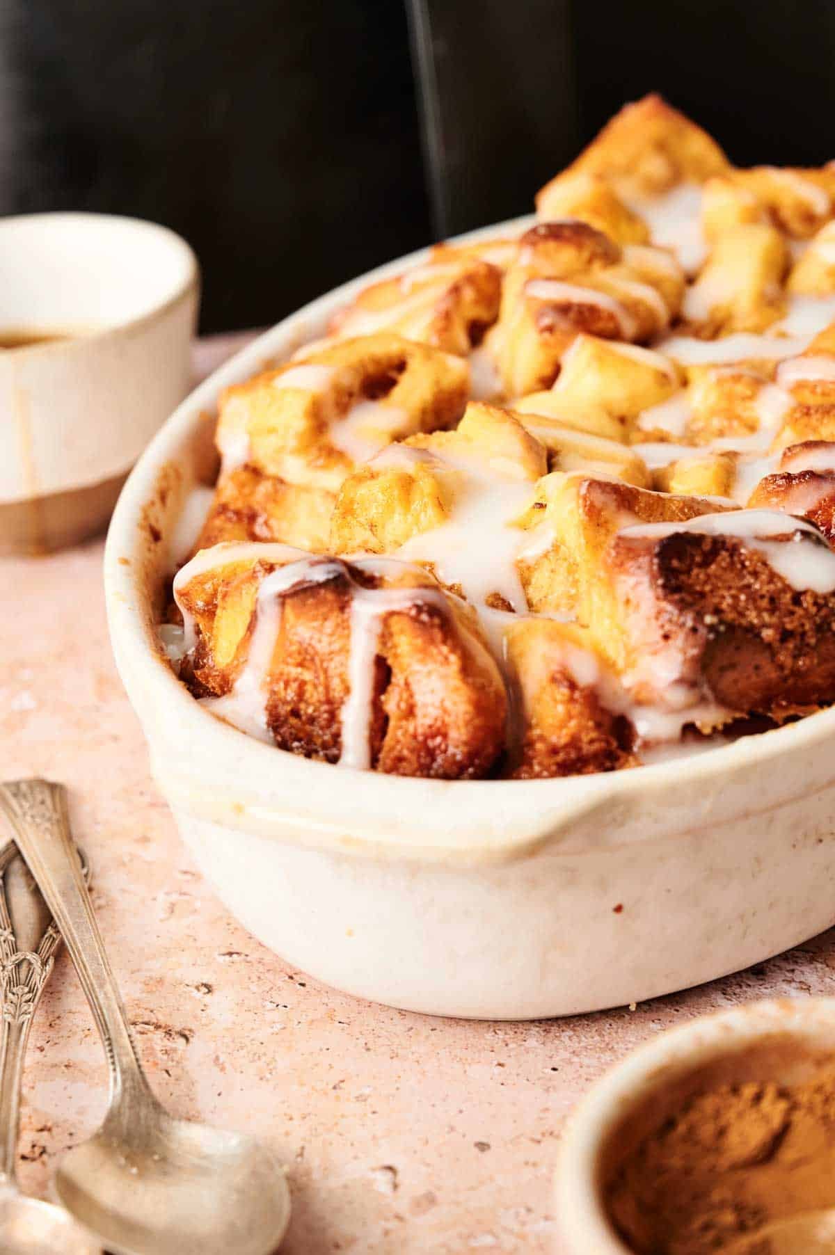 A white baking dish filled with Air Fryer Cinnamon Roll Casserole topped with white icing, placed on a light countertop with a small bowl and spoons nearby.