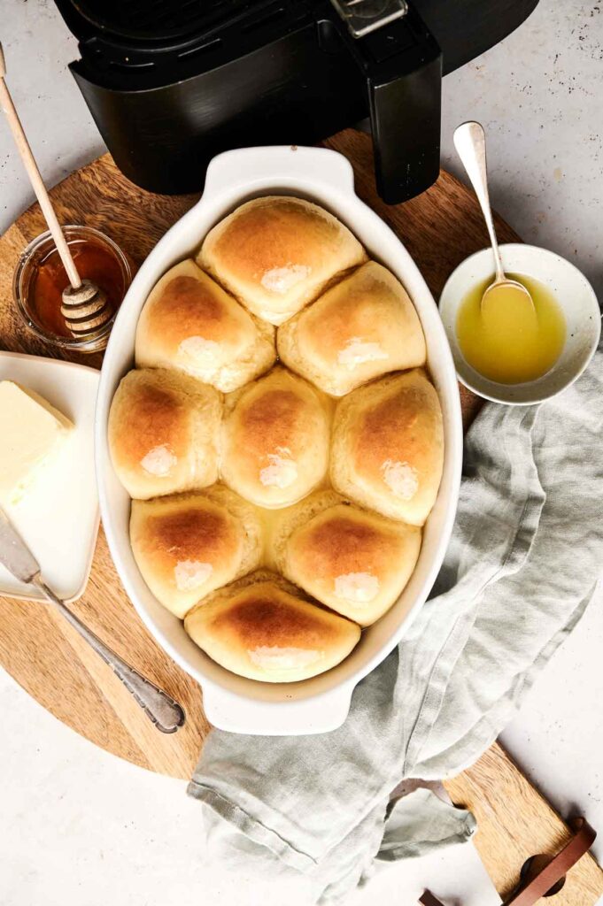 A white oval dish with eight golden-brown Air Fryer Dinner Rolls, next to a bowl of butter, a bowl of honey with a dipper, and a gray cloth on a wooden board.