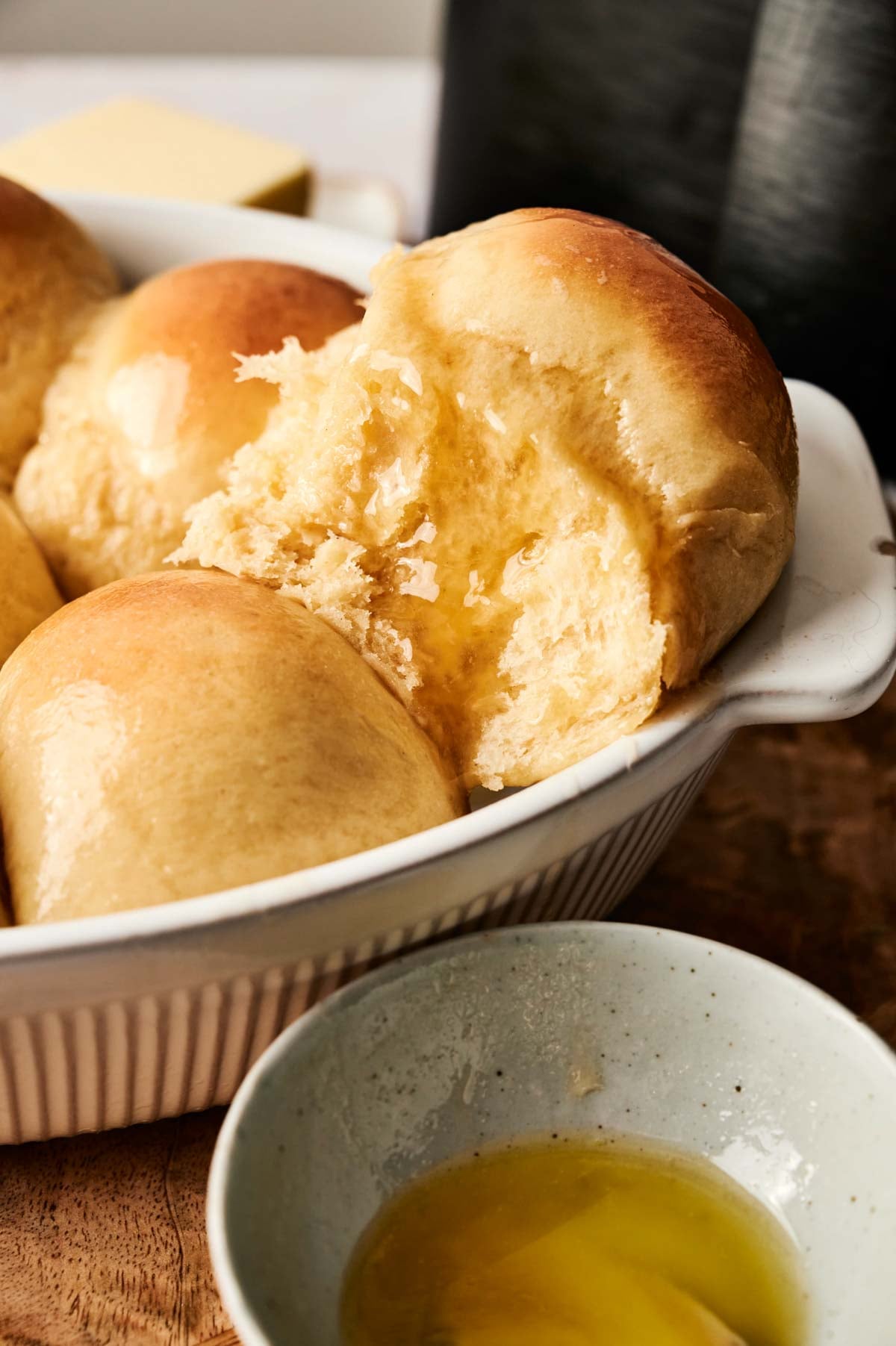 A close-up of golden brown Air Fryer Dinner Rolls in a white baking dish, with one roll partially torn open, next to a small bowl containing melted butter.
