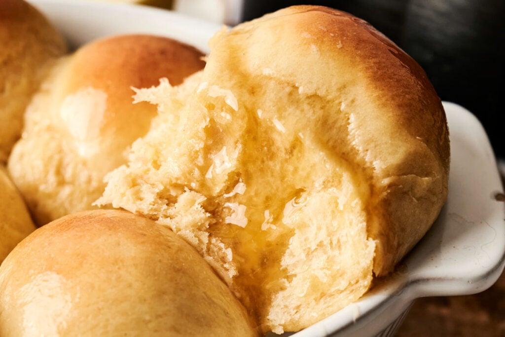 Close-up of soft, golden-brown Air Fryer Dinner Rolls in a white dish, with one roll pulled apart to reveal a fluffy interior and a drizzle of melted butter.