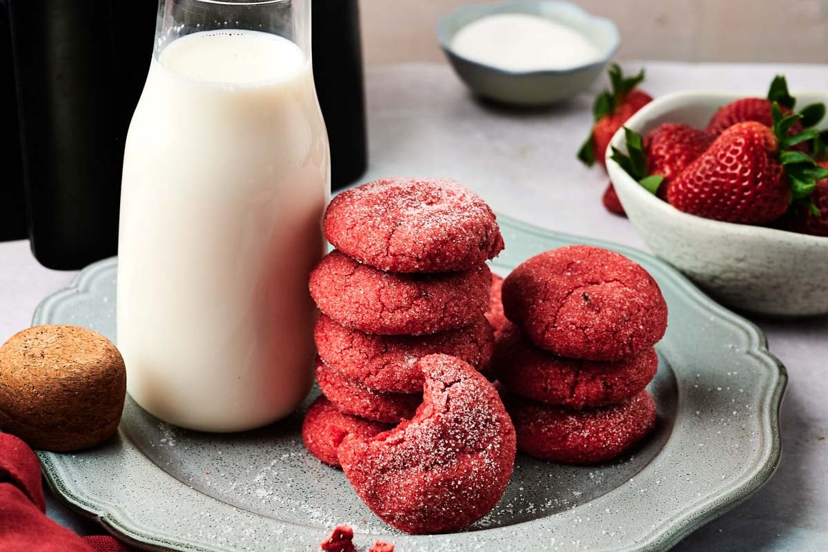 A plate with stacked Air Fryer Strawberry Cookies, a bitten cookie in front, a glass bottle of milk, and bowls of strawberries and sugar in the background.