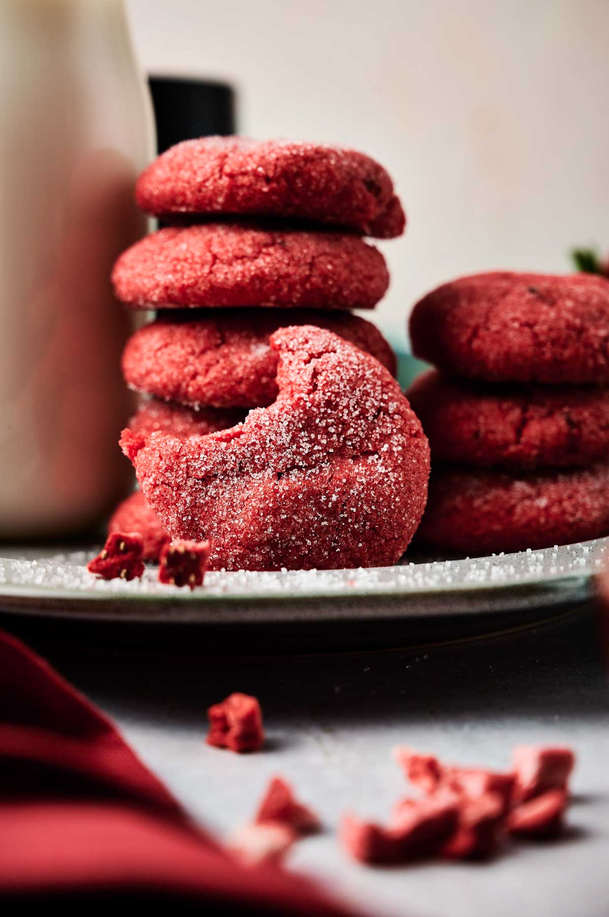A plate of red Air Fryer Strawberry Cookies, with one cookie in front showing a bite taken out of it. Crumbs are scattered nearby.