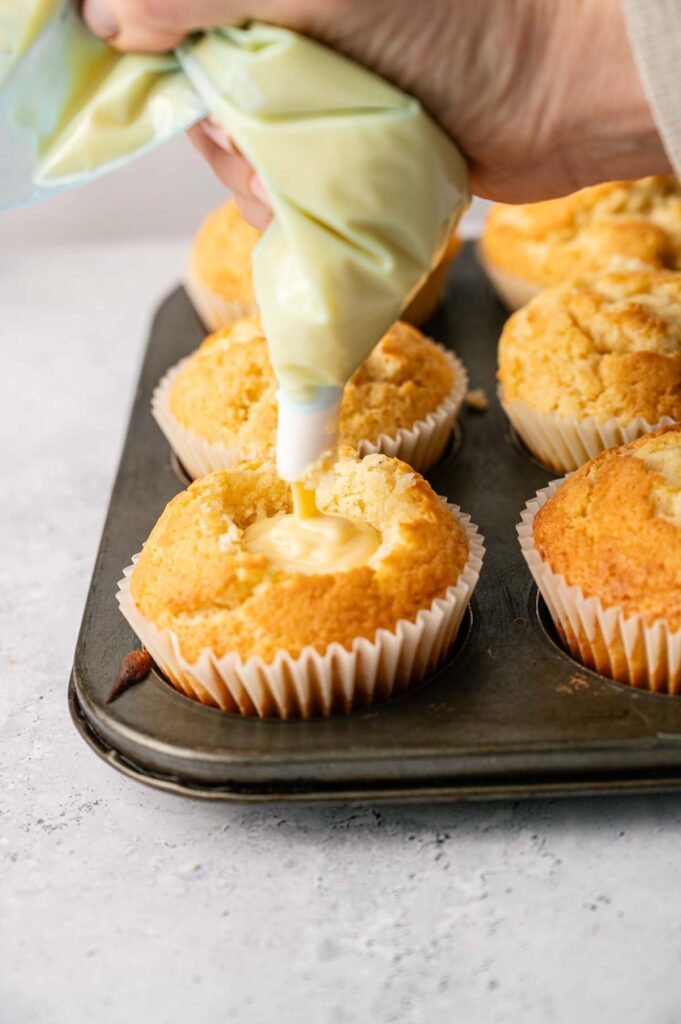 A hand uses a piping bag to fill Boston Cream Pie Cupcakes with cream filling while several cupcakes sit in a muffin tray.