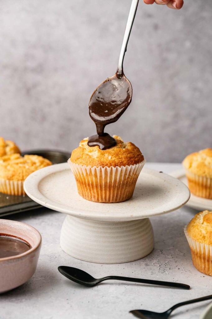 A hand holds a spoon dripping chocolate sauce onto a plain muffin on a stand, evoking the classic flavors of Boston Cream Pie Cupcakes. More muffins and utensils are visible in the background.