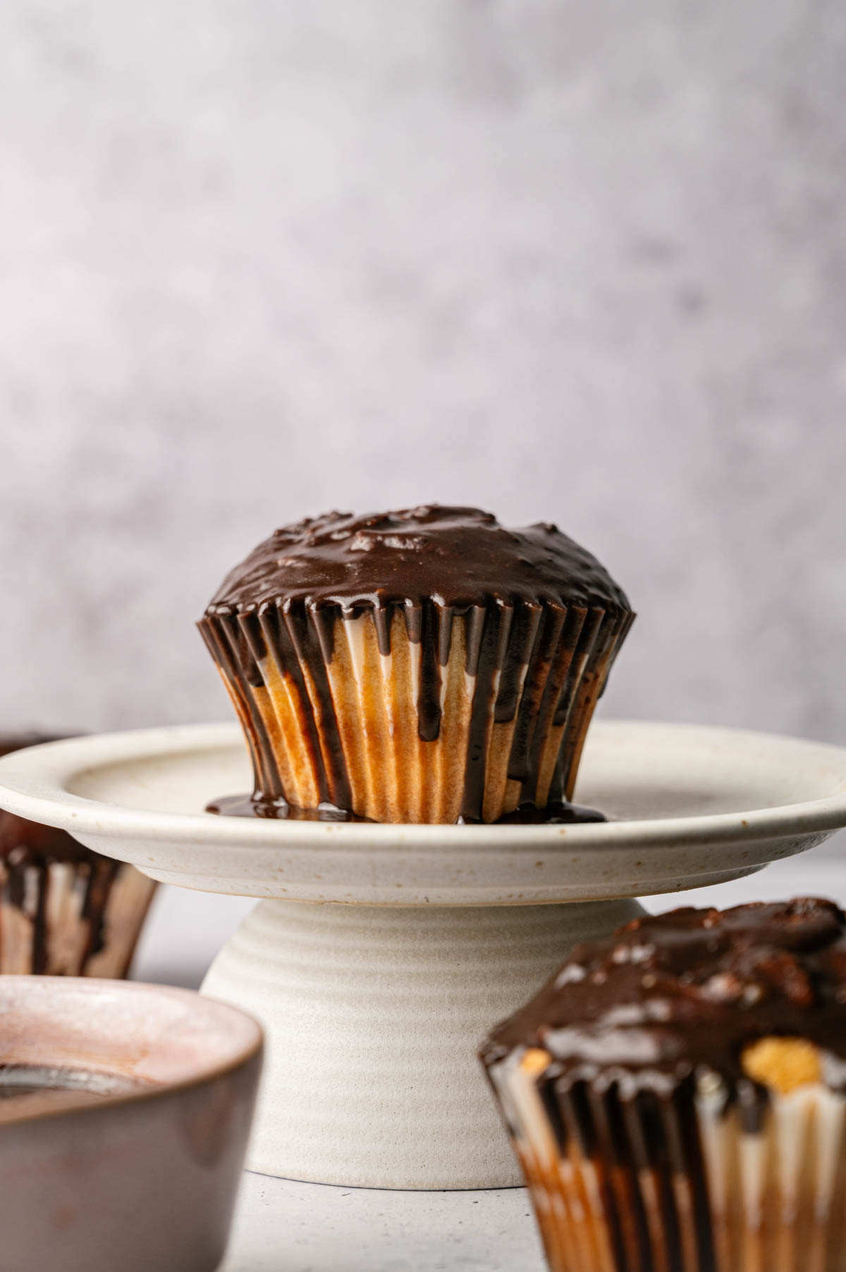 A single Boston Cream Pie Cupcake with chocolate frosting sits on a white pedestal plate, with another partially visible cupcake in the foreground.