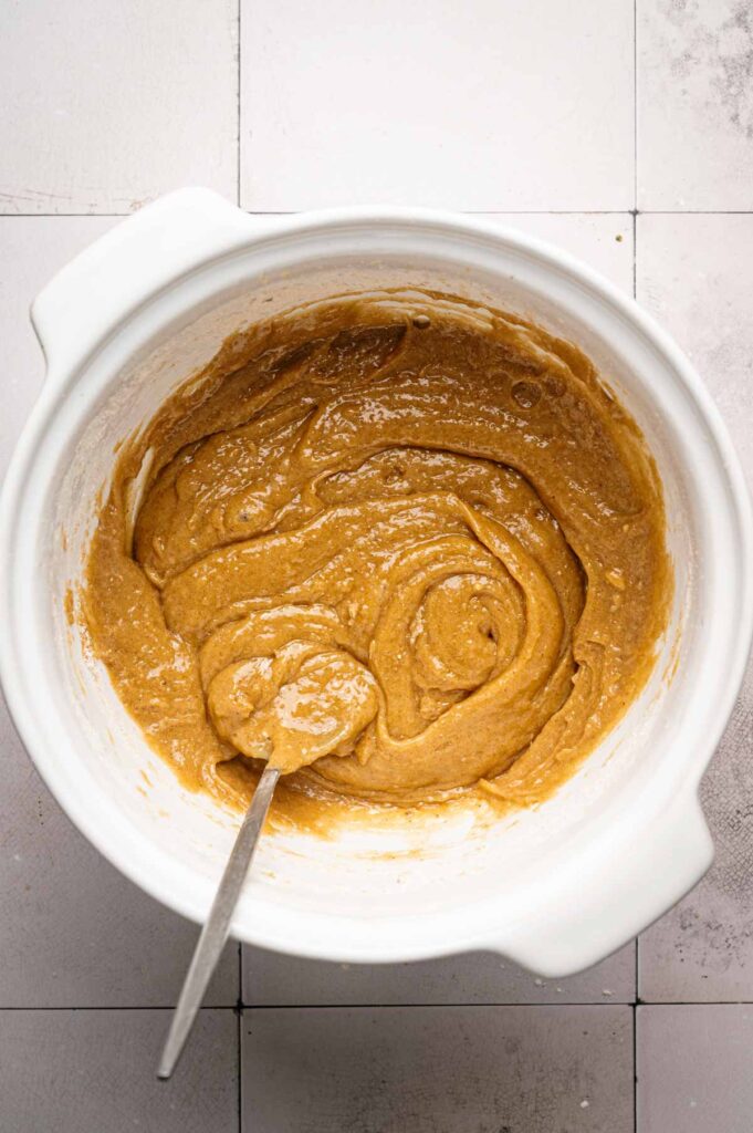 A white mixing bowl filled with light brown butter pecan cookie batter is shown with a metal spoon resting inside. The bowl sits on a tiled countertop.