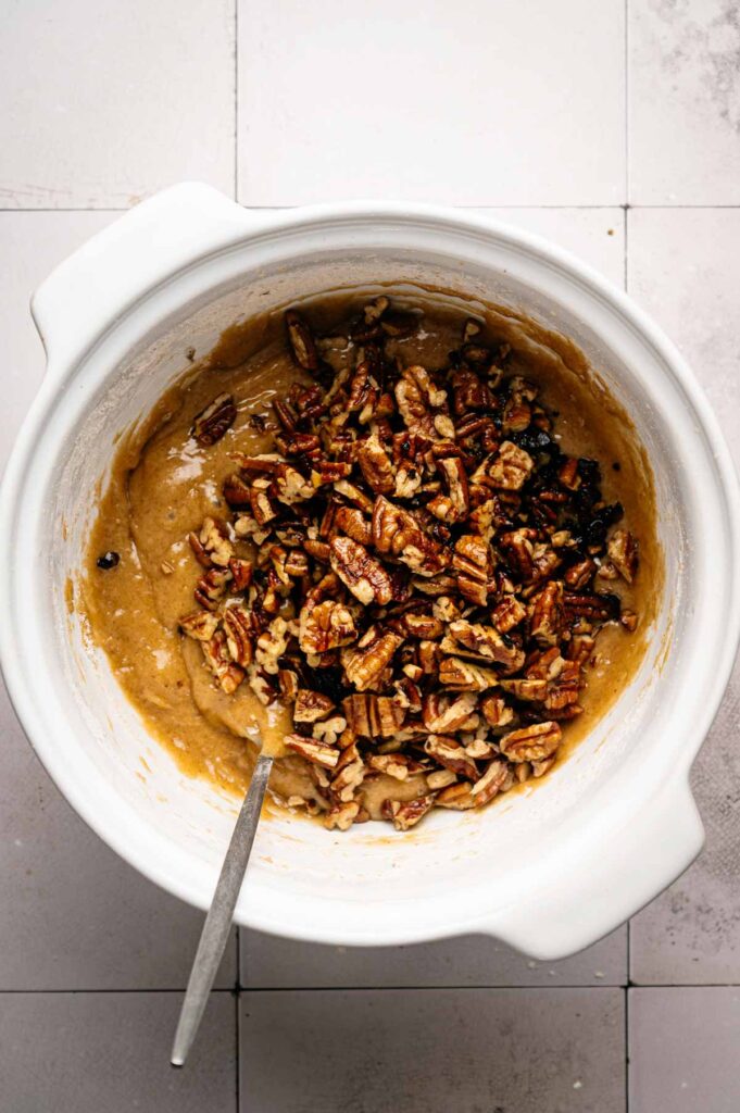 White mixing bowl with light brown batter and chopped pecans being stirred with a metal spoon, sitting on a tiled surface—the perfect start for delicious Butter Pecan Cookies.