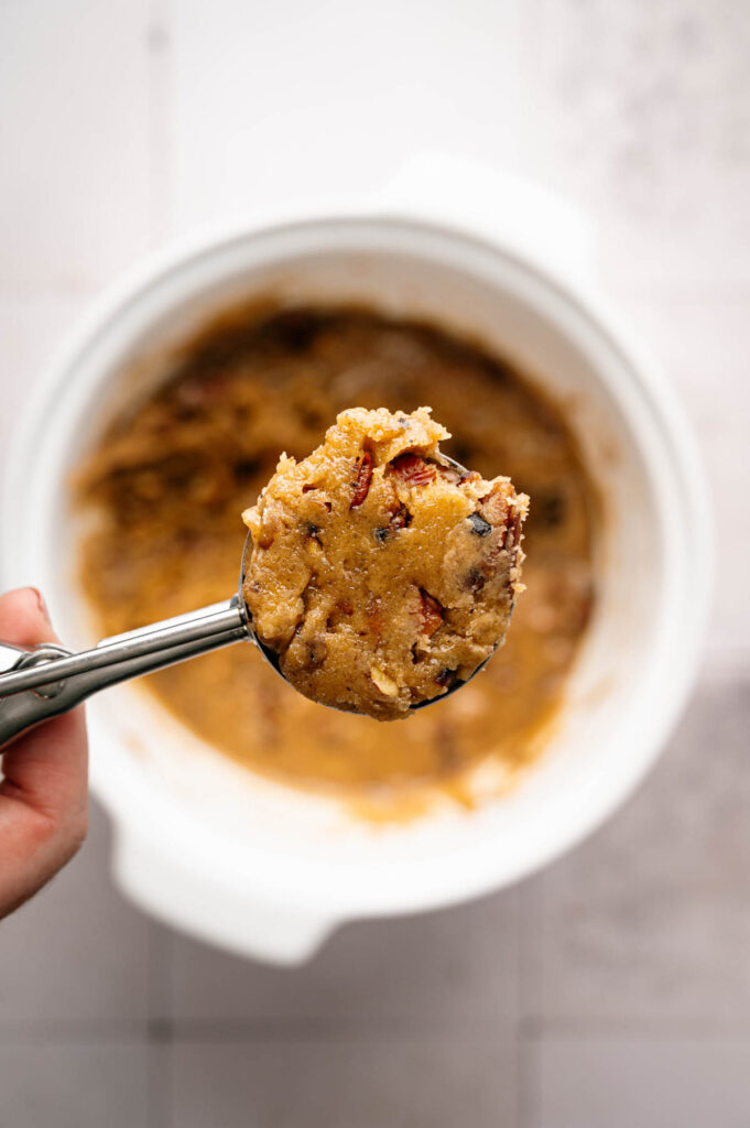 A hand holds a metal cookie scoop filled with raw Butter Pecan Cookies dough above a white mixing bowl containing more dough.