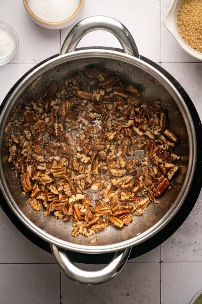 Chopped pecans being toasted in a stainless steel pan for Butter Pecan Cookies, with bowls of sugar and other ingredients visible nearby on a tiled surface.