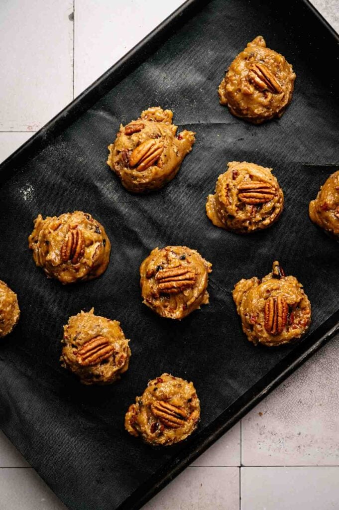 A baking tray lined with parchment paper holds unbaked Butter Pecan Cookies, each scoop of dough adorned with a pecan half.